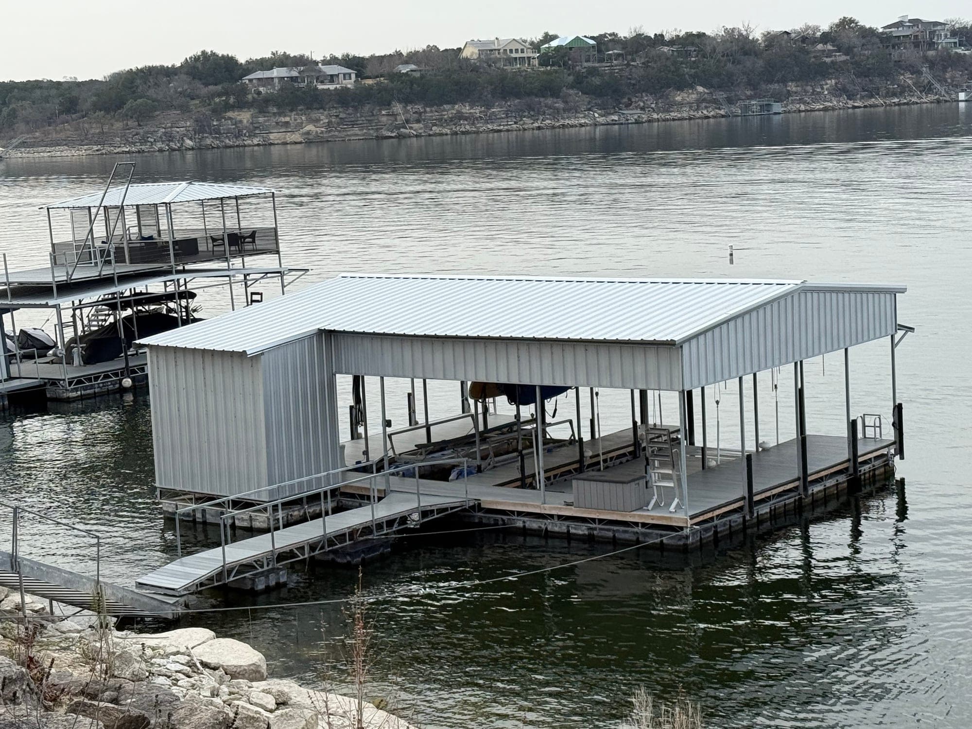 Finished two-slip boat dock on Lake Travis with a white metal roof, an upper sundeck and a walkway out from the rocky bank.